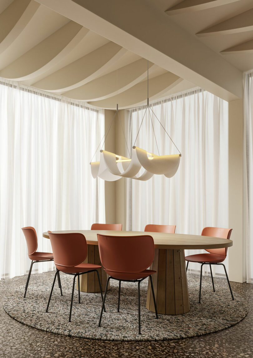 A modern dining area features a round wooden table, six red chairs, and a textured rug. Enhancing the room's elegance are white drapes paired with three unique pendant lights gracefully illuminating the space.