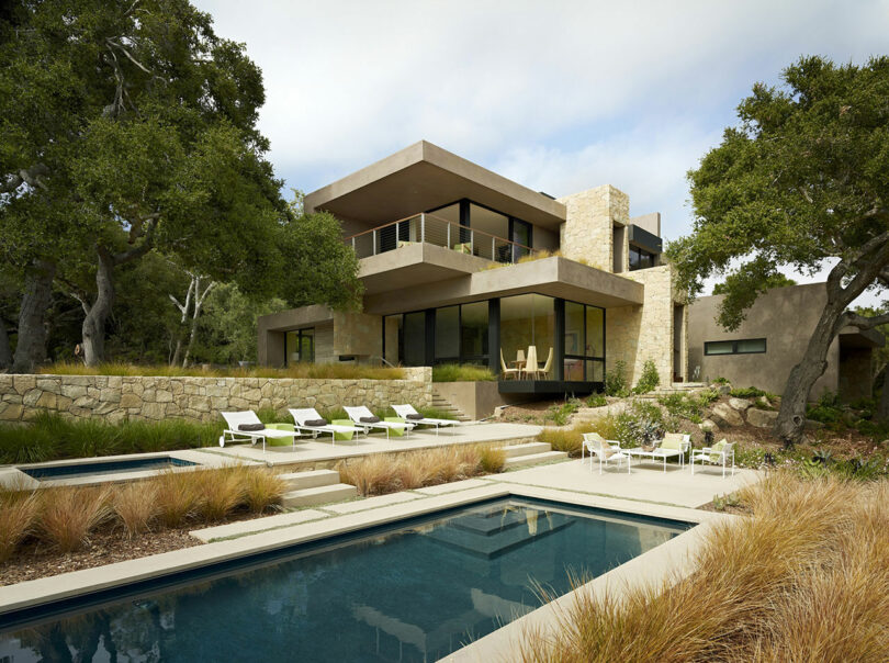 Modern two-story house with stone and glass elements, designed by Ron Radziner, surrounded by trees. In front, a landscaped yard has a pool area with lounge chairs and an outdoor seating space.