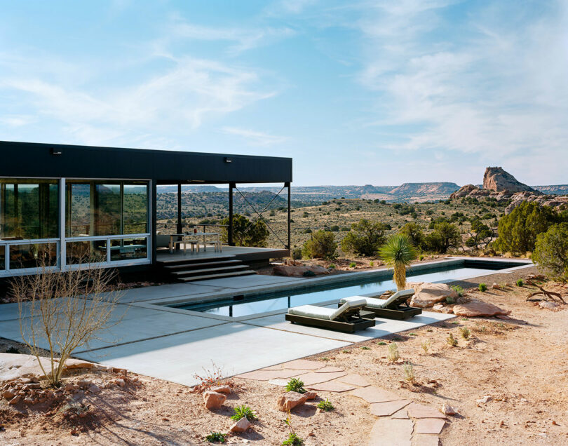 A modern glass house designed by Ron Radziner, featuring a patio and pool, is situated in a desert landscape with distant mountains. The pool area includes lounge chairs, and minimal vegetation surrounds the property.