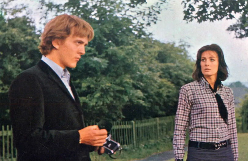 A man in a black jacket holds a camera and stands next to a woman in a plaid shirt, both outdoors with trees and a fence in the background.