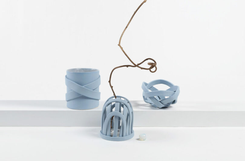 Three blue ceramic objects are displayed on a white surface: a cylindrical vase, a circular decorative item with a twig, and a spherical sculpture. A small white pebble is placed to the right.