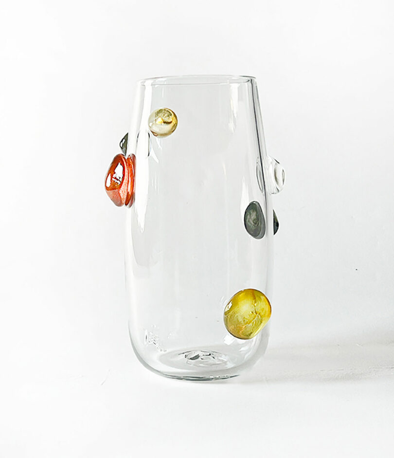 A clear glass vase by Jess Humphrey with round, colorful decorative elements attached to its surface. The elements are red, yellow, green, and black. The vase is placed against a white background.