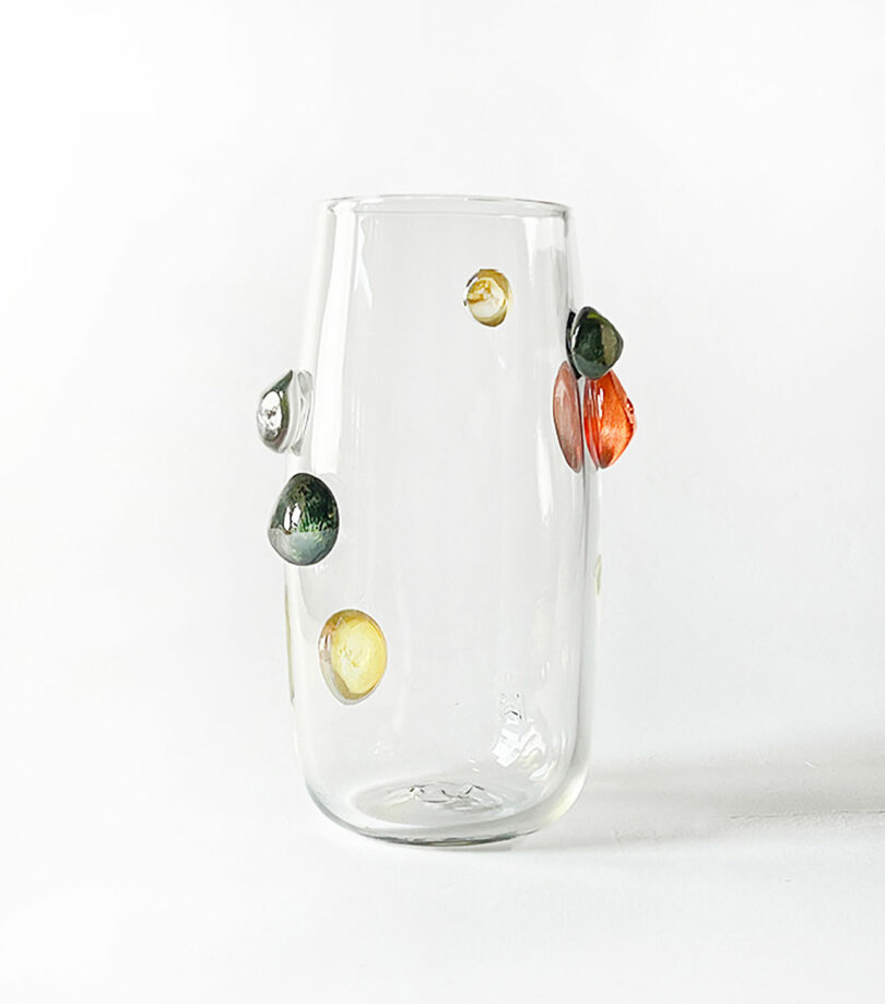 A clear glass vase by Jess Humphrey with multiple colorful spherical embellishments on its surface against a plain white background.
