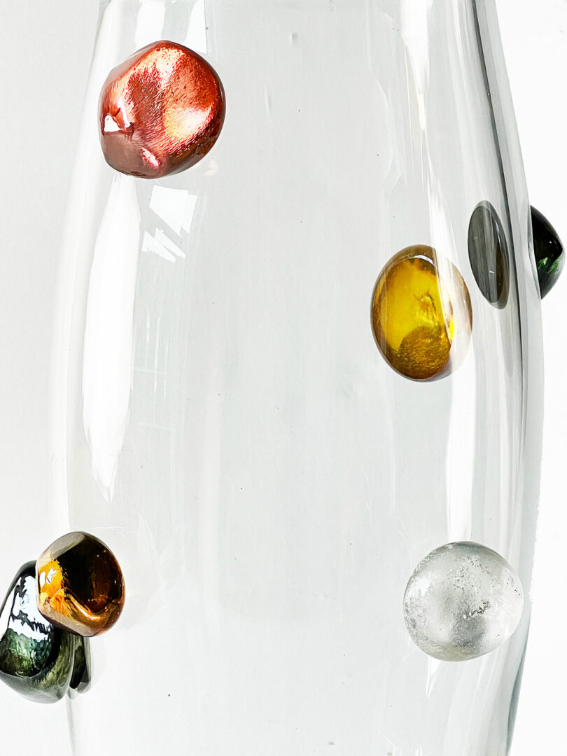 A clear glass vase by Jess Humphrey with four small, colorful orbs embedded on its surface, including red, amber, green, and clear.