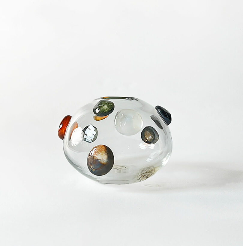 A clear glass vase by Jess Humphrey with colorful spherical shapes embedded on its surface, placed against a plain white background.