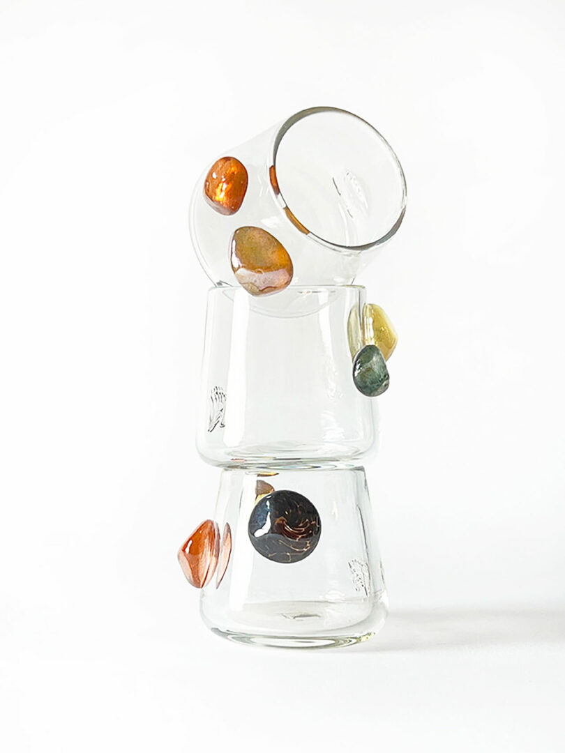 Three stacked clear glasses by Jess Humphrey adorned with colorful, irregularly shaped stones against a white background.