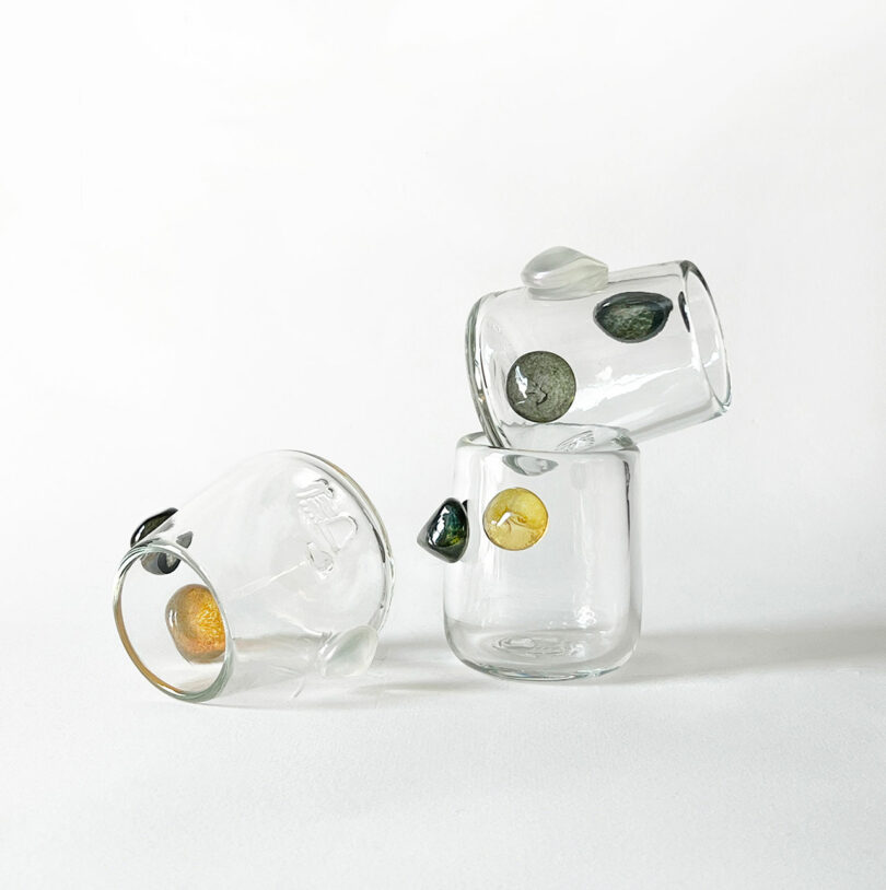 Three clear glass cups by Jess Humphrey with embedded colored stones, arranged in a minimalist composition against a white background. Two cups are stacked, one is tipped over.