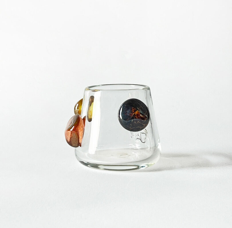 A clear glass tumbler by Jess Humphrey with three decorative elements in amber, yellow, and copper attached to its surface is placed against a plain white background.