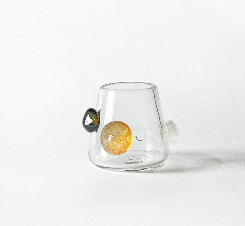 A glass tumbler by Jess Humphrey with a conical shape, featuring protruding decorative elements in orange, black, and white on its exterior.