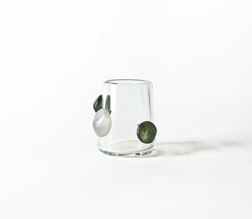 A clear glass tumbler by Jess Humphrey with three large, irregularly shaped stones attached to its exterior surface, set against a white background.
