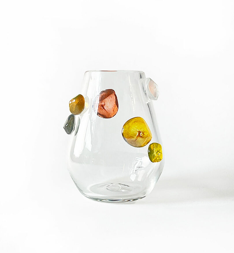 A clear glass vase by Jess Humphrey featuring irregularly-shaped, colorful accents attached to its surface.