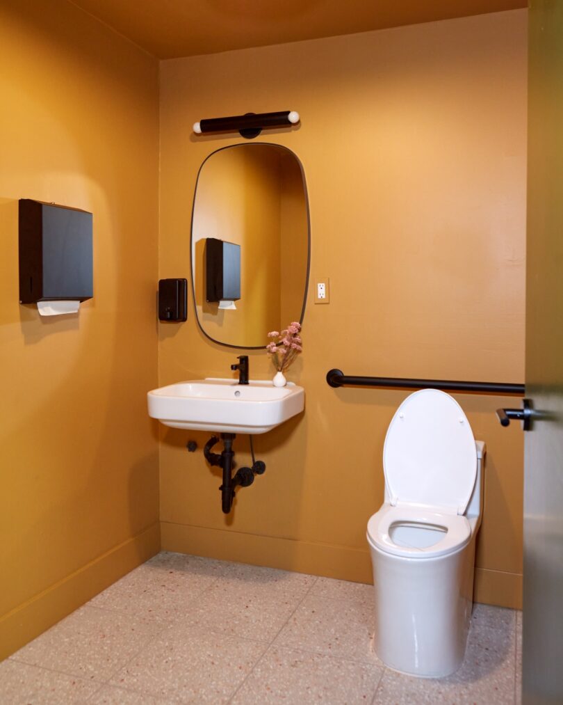 A small bathroom with mustard yellow walls, featuring a white wall-mounted sink with a mirror above, soap dispenser, towel holder, and a toilet with grab bars next to it
