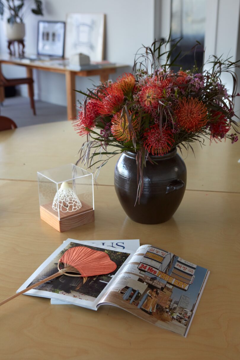 A vase of vibrant flowers, an open magazine, and a decorative object in a glass case are arranged on a wooden table. Framed pictures are visible in the background