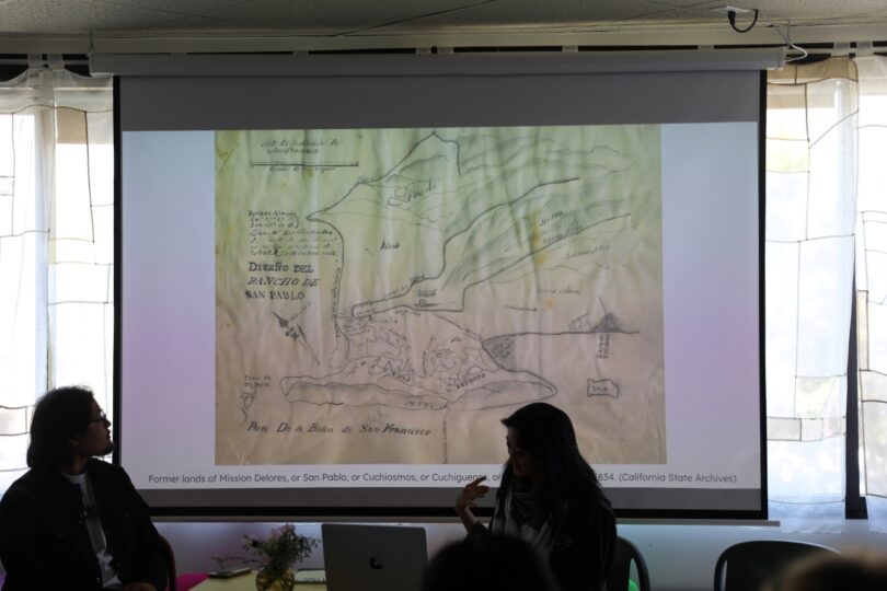 Two people are seated in front of a projected image of a historical map. The map is labeled to show the former lands of Mission Dolores and areas around San Francisco in 1834