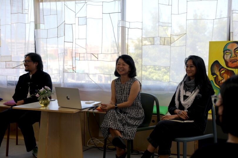 Three people sit at a panel discussion in a room with large windows. One person has a laptop and flowers on a small table. A colorful painting is displayed in the background