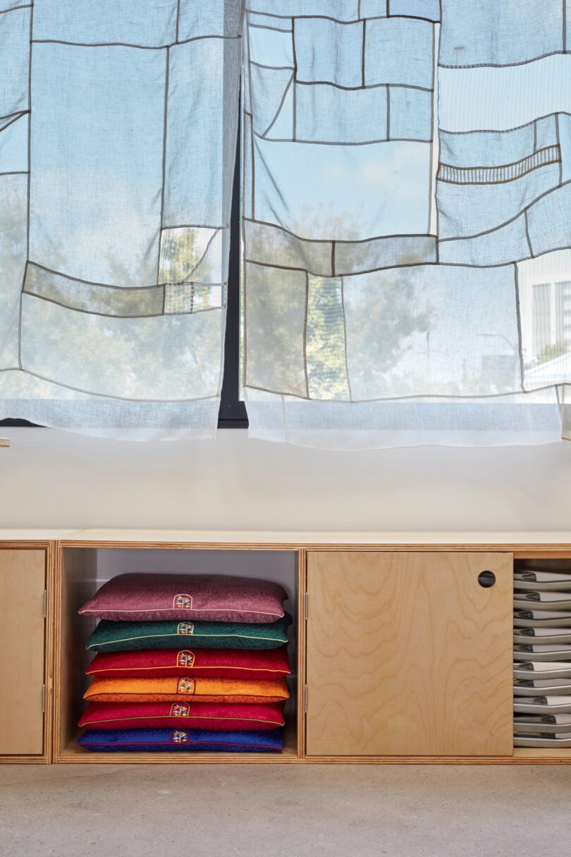 A stack of colorful cushions is stored in a wooden cabinet beneath a low bench seat. Sunlight filters through patterned window curtains, casting a bright, airy atmosphere