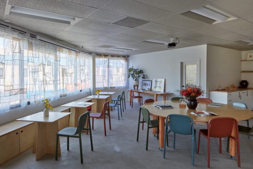 An office with several round and rectangular tables, colored chairs, large windows with sheer curtains, and various plants and books around the room.
