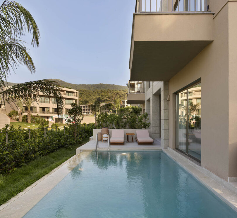 A serene pool beside a modern building with lounge chairs, greenery, and distant mountains in the background.