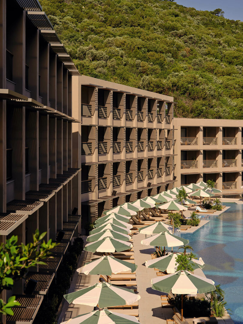Poolside view of a hotel with multiple balconies, sun loungers, and umbrellas overlooking a serene pool. Lush green hillside in the background.