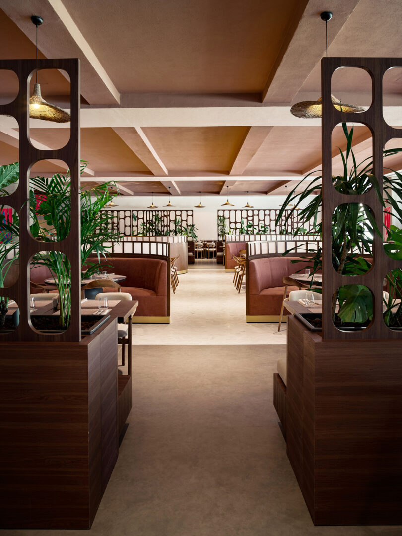 Modern restaurant interior with wooden partitions, brown seating, and green plants. Tables are neatly set, and pendant lights hang from the ceiling.