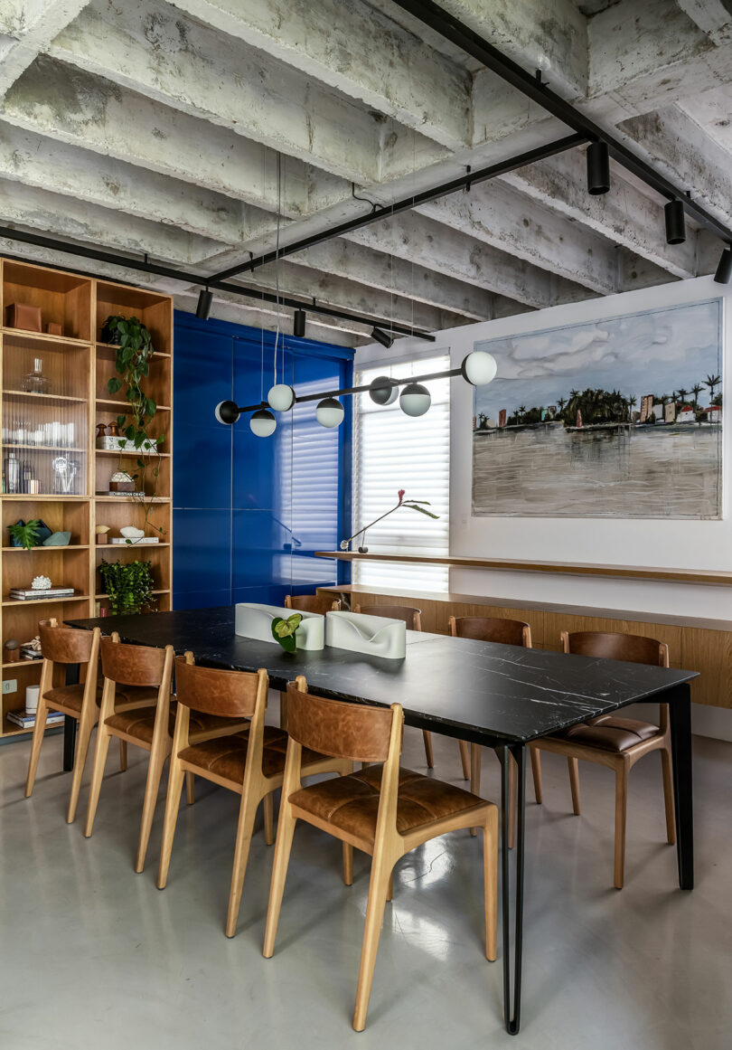 A modern dining room with a black table, eight wooden chairs, a blue accent wall, a shelf with plants, and a large painting. Exposed concrete ceiling with track lighting and decorative pendant lights.