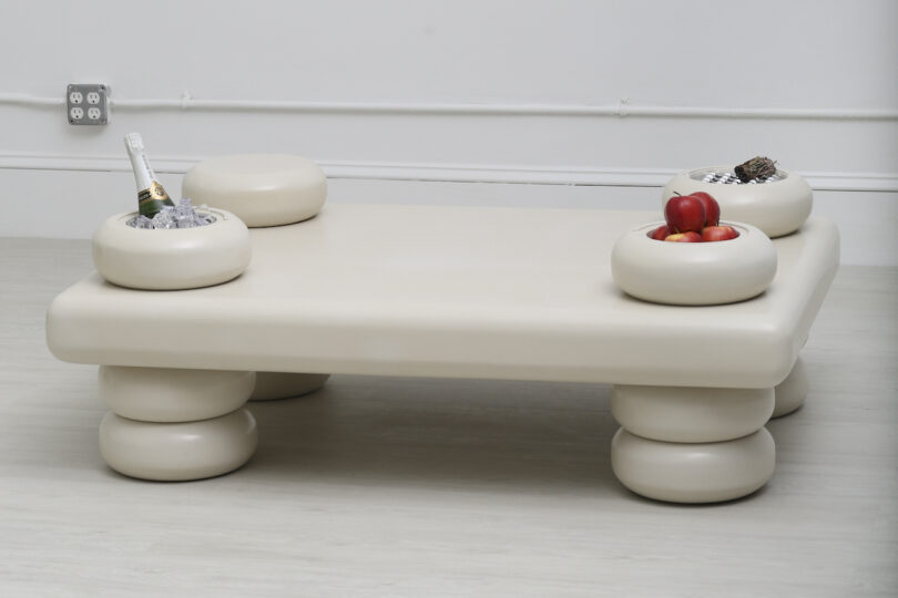A beige, uniquely designed coffee table with cylindrical legs and round storage compartments, holding a bottle in ice and fruits, is set in a minimalist room with white floor and walls