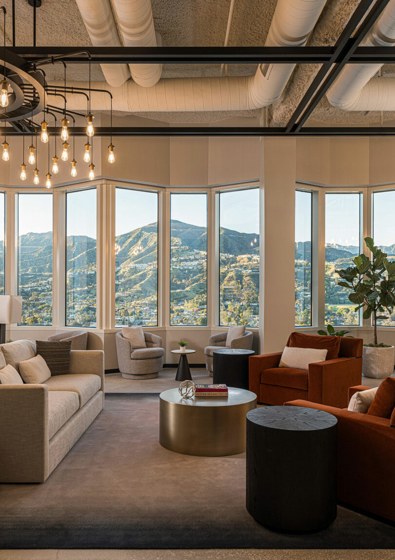 A modern lounge area with large windows offering scenic mountain views. The space features comfortable seating with sofas and armchairs, a central round coffee table, and overhead hanging lights.