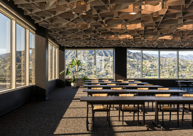 An empty conference room with large windows showcasing a mountain view, rows of tables and chairs, a geometric ceiling design, and a plant by the window.