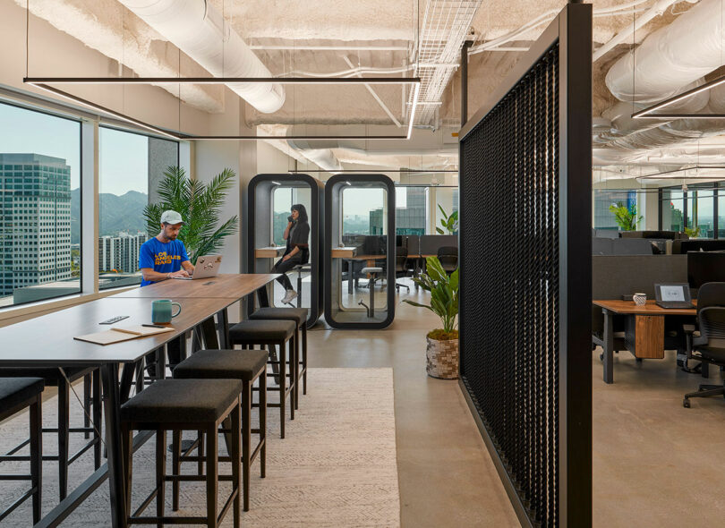 Modern office space with open seating, individual work booths, and large windows providing natural light. One person works on a laptop at a communal table, while another is seated in a private booth.