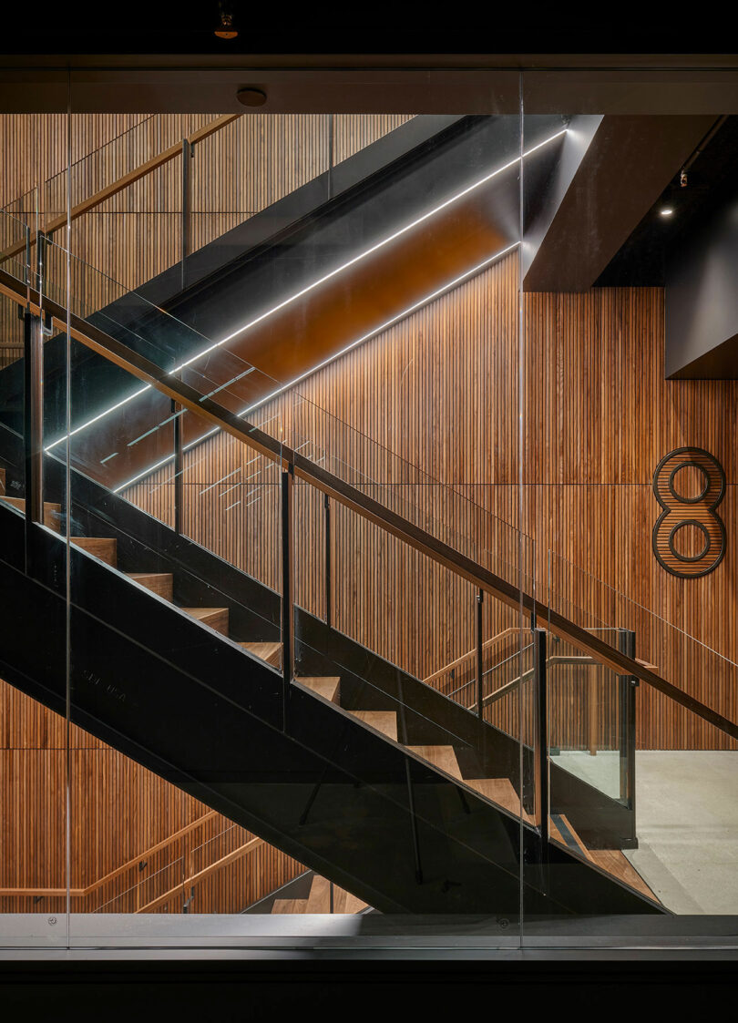 A modern interior staircase with black steps and glass railings ascends between wooden slatted walls and an illuminated number eight on the right.