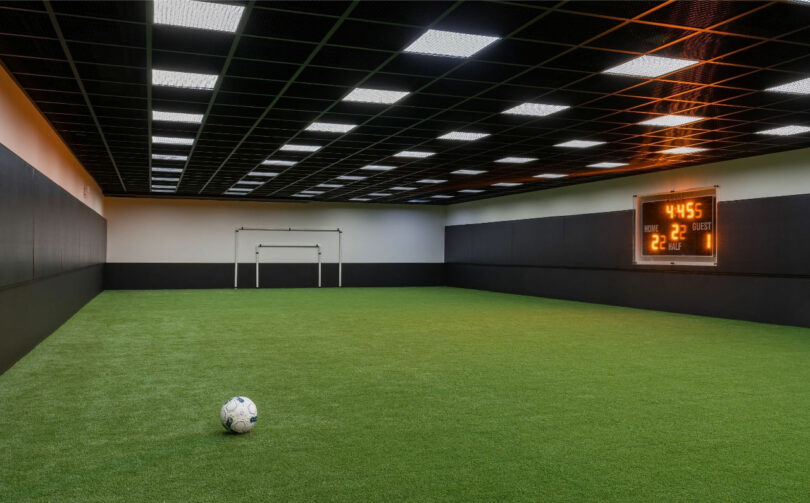 Indoor soccer field with artificial turf, a lone soccer ball, and a scoreboard indicating a home team score of 22 and guest team score of 28 with 4:55 remaining.