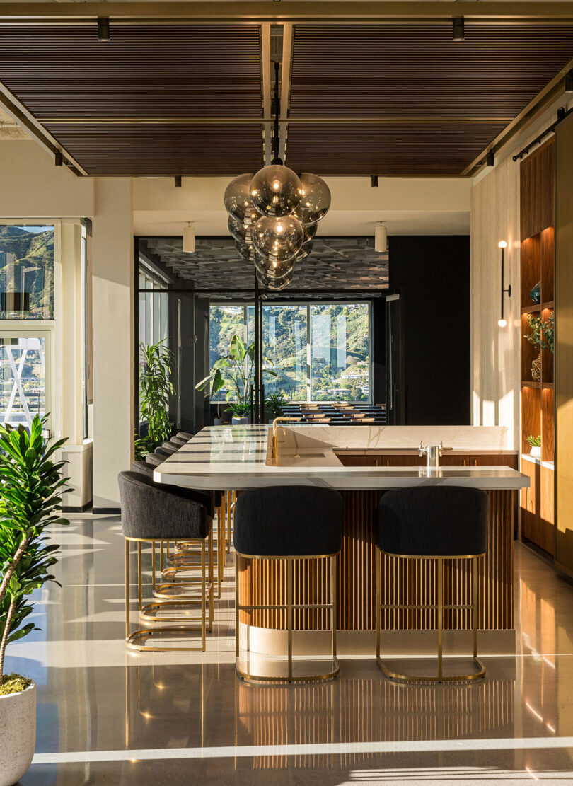 A modern kitchen with a central island, stools, hanging light fixtures, and large windows providing natural light. The area is decorated with plants and has a polished floor.