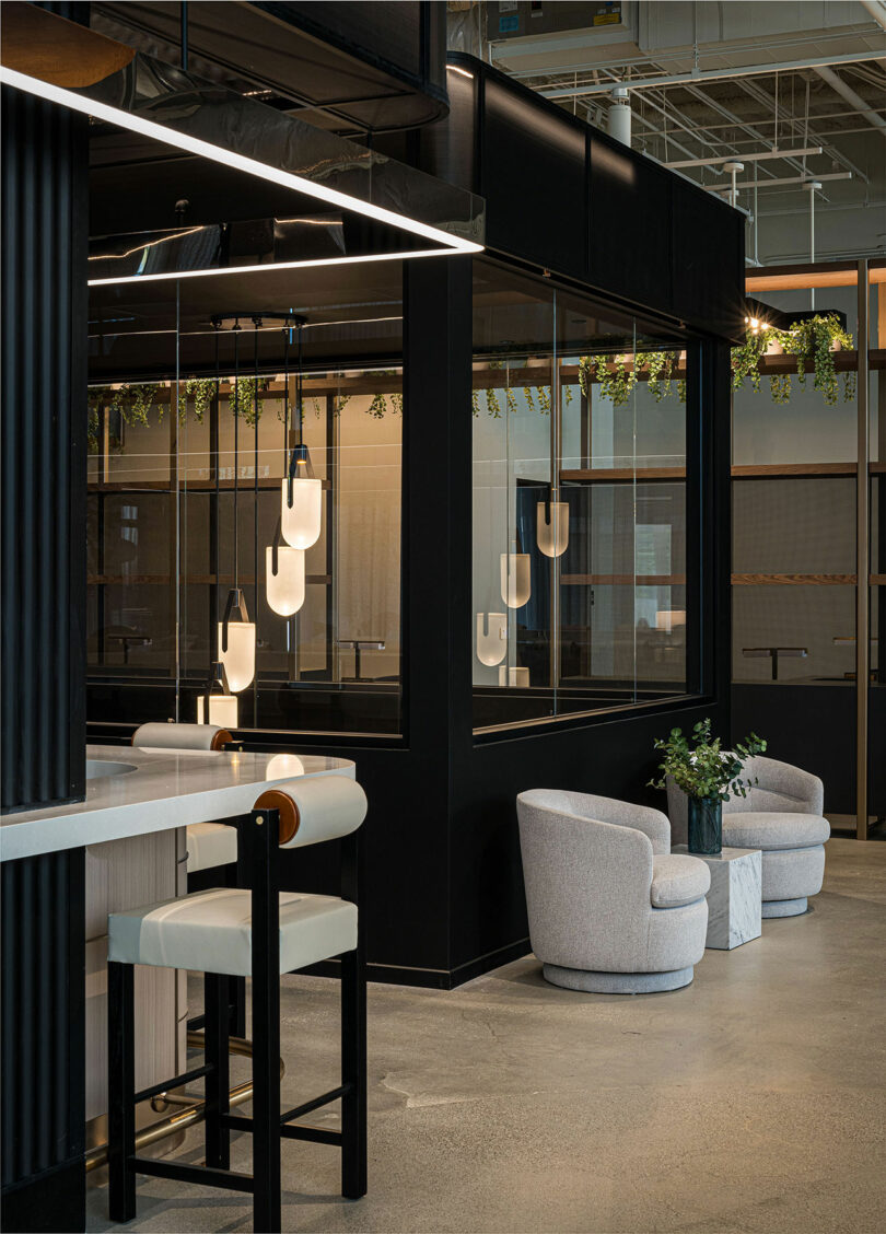 Modern interior space with sleek black and white furniture, including a bar stool and armchairs, pendant lights, and green plants. Large windows and minimalistic design elements are present.
