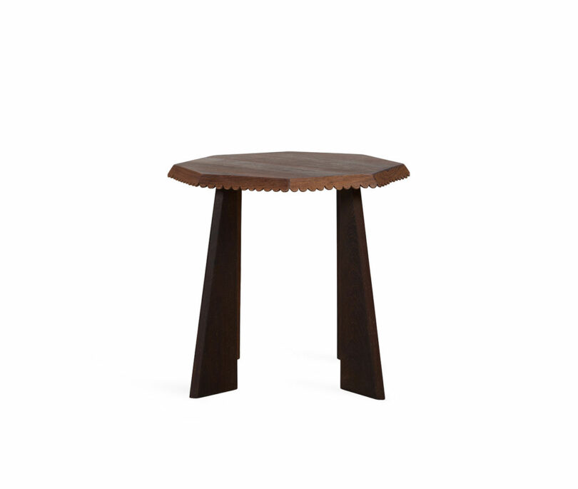 A wooden three-legged table with an octagonal top and scalloped edge detailing stands against a plain white background