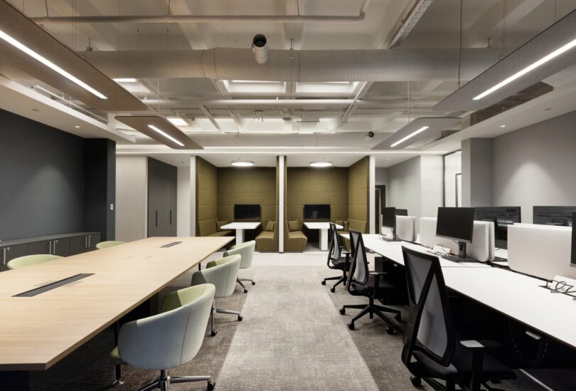 A modern office space with long wooden desks, ergonomic chairs, computer workstations, recessed ceiling lights, and partition areas with individual workspaces.