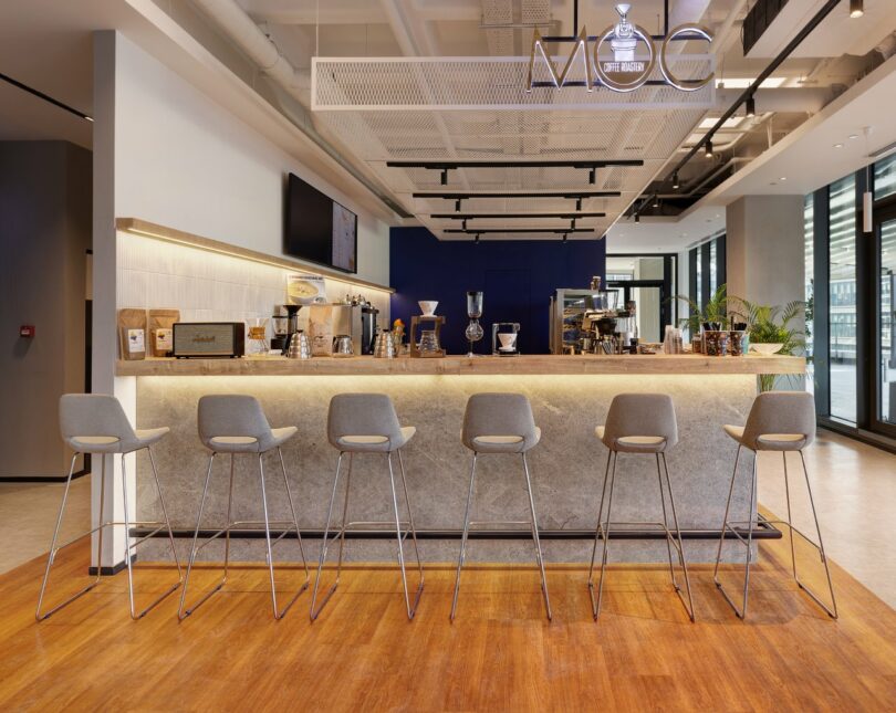 A modern coffee shop with a long counter, high stools, coffee brewing equipment, and a sign reading "MDC." The space features wooden flooring, a blue accent wall, and large windows.