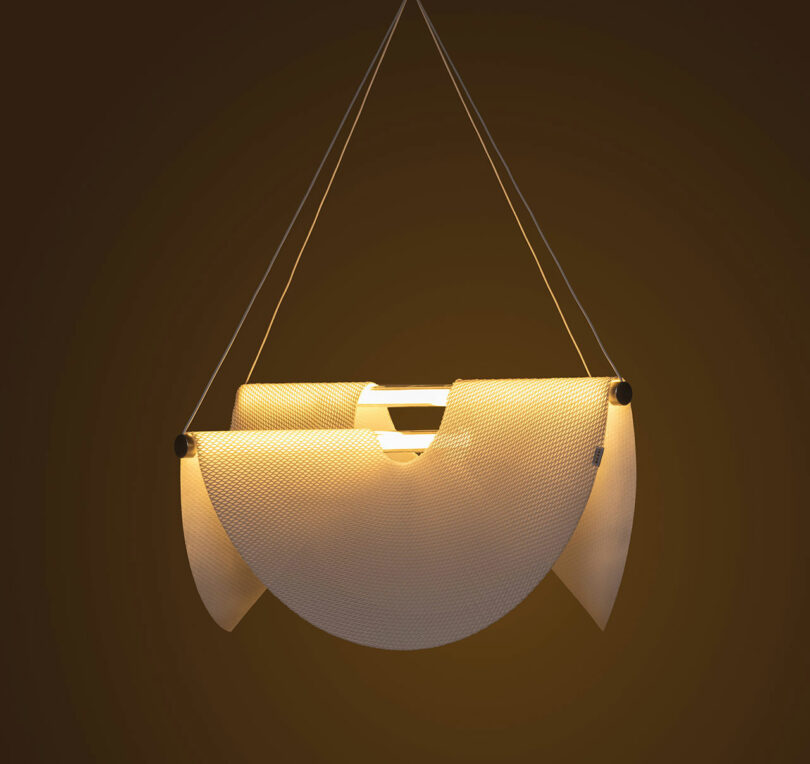 The Drape Light by Jamie Wolfond with three curved, overlapping white panels emitting a warm glow, suspended by thin cables.