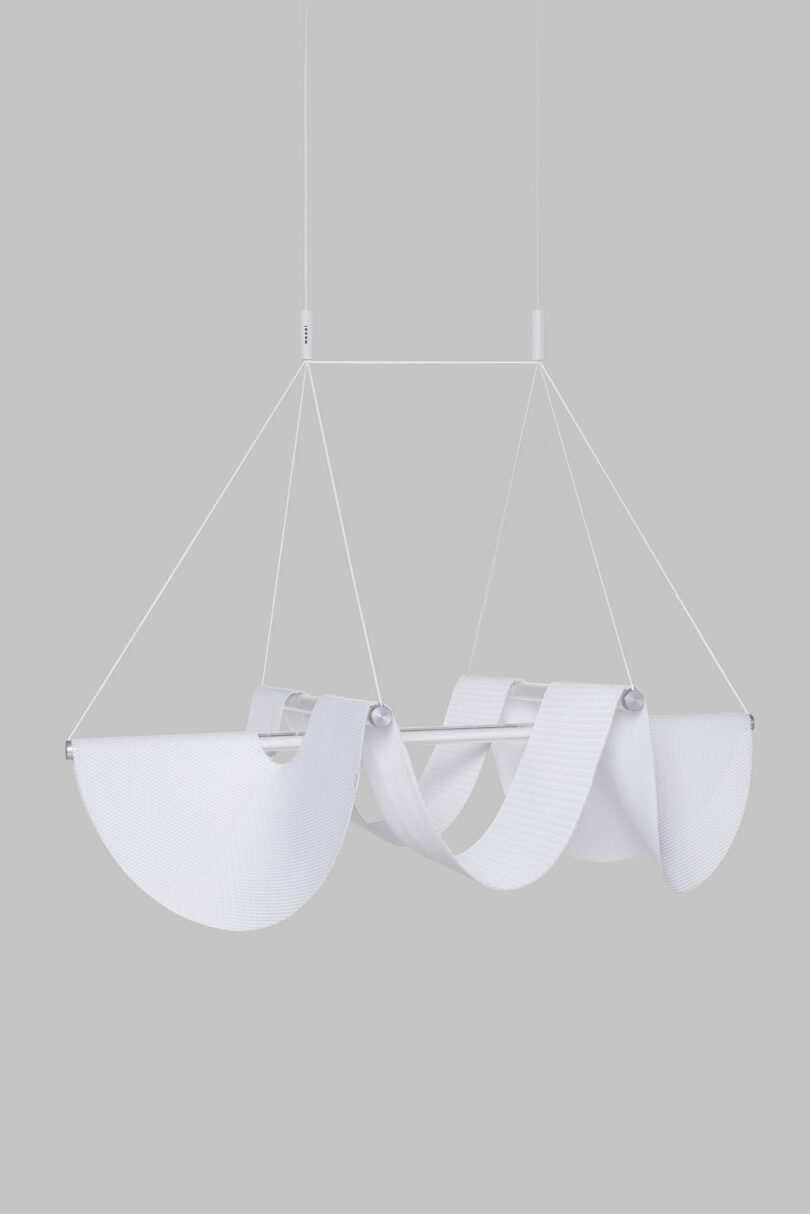 The Drape Light by Jamie Wolfond, with an abstract, wave-like design, suspended by thin wires from a ceiling against a plain gray background.