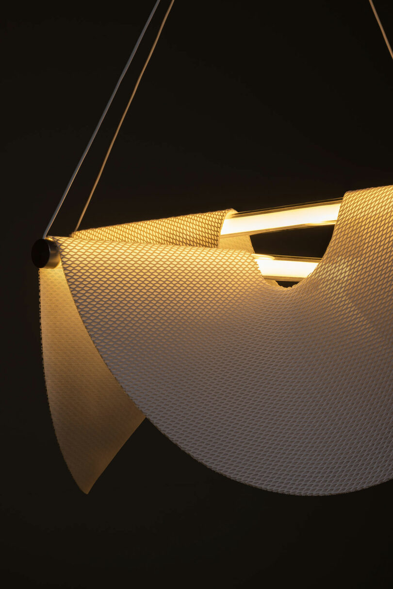 The Drape Light by Jamie Wolfond with a white perforated curved shade and two exposed tubular bulbs, suspended by two thin cords. The light is turned on in a dark background.