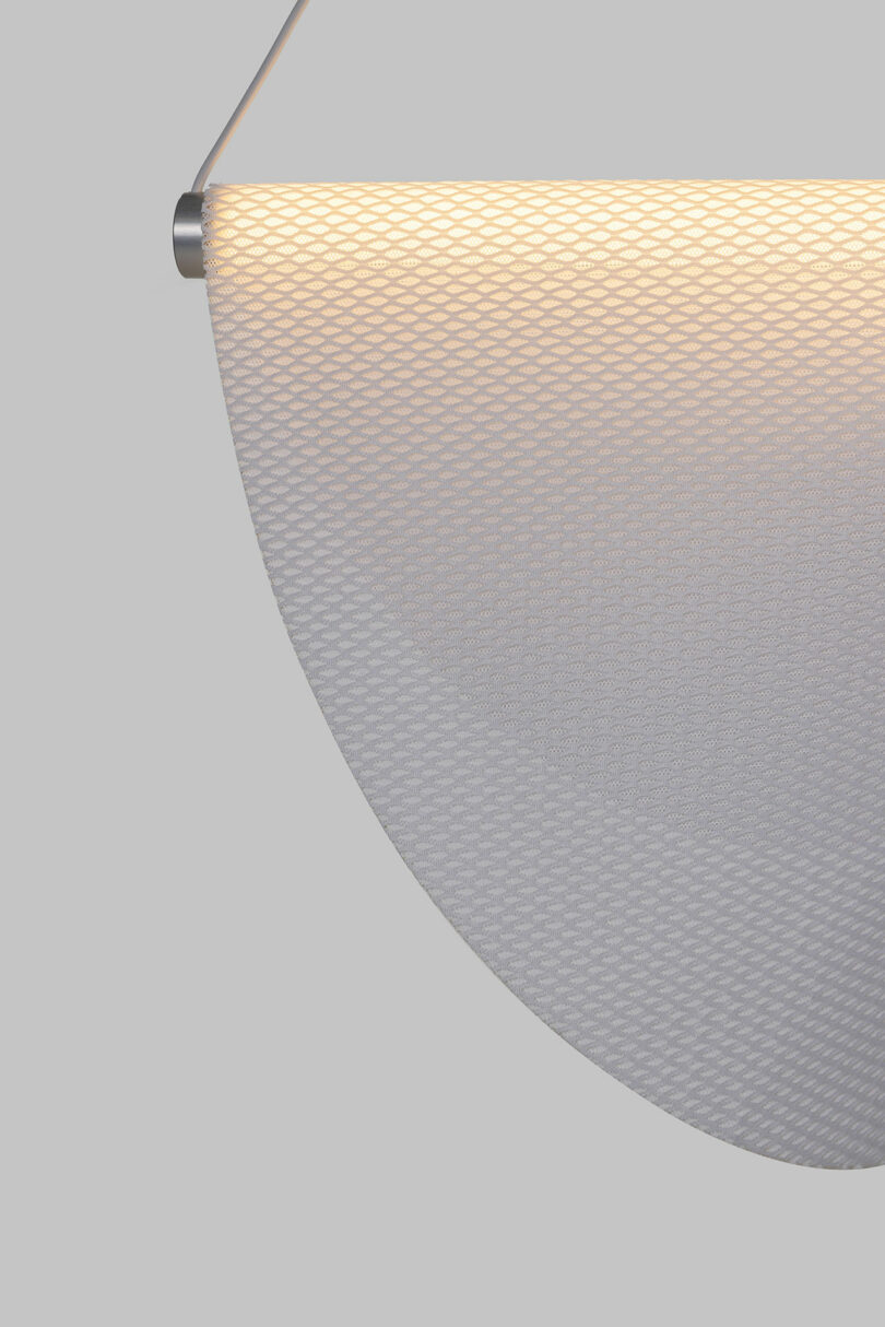 Close-up view of the Drape Light by Jamie Wolfond with a mesh design, emitting a soft glow. Hanging from a metallic rod against a plain gray background.