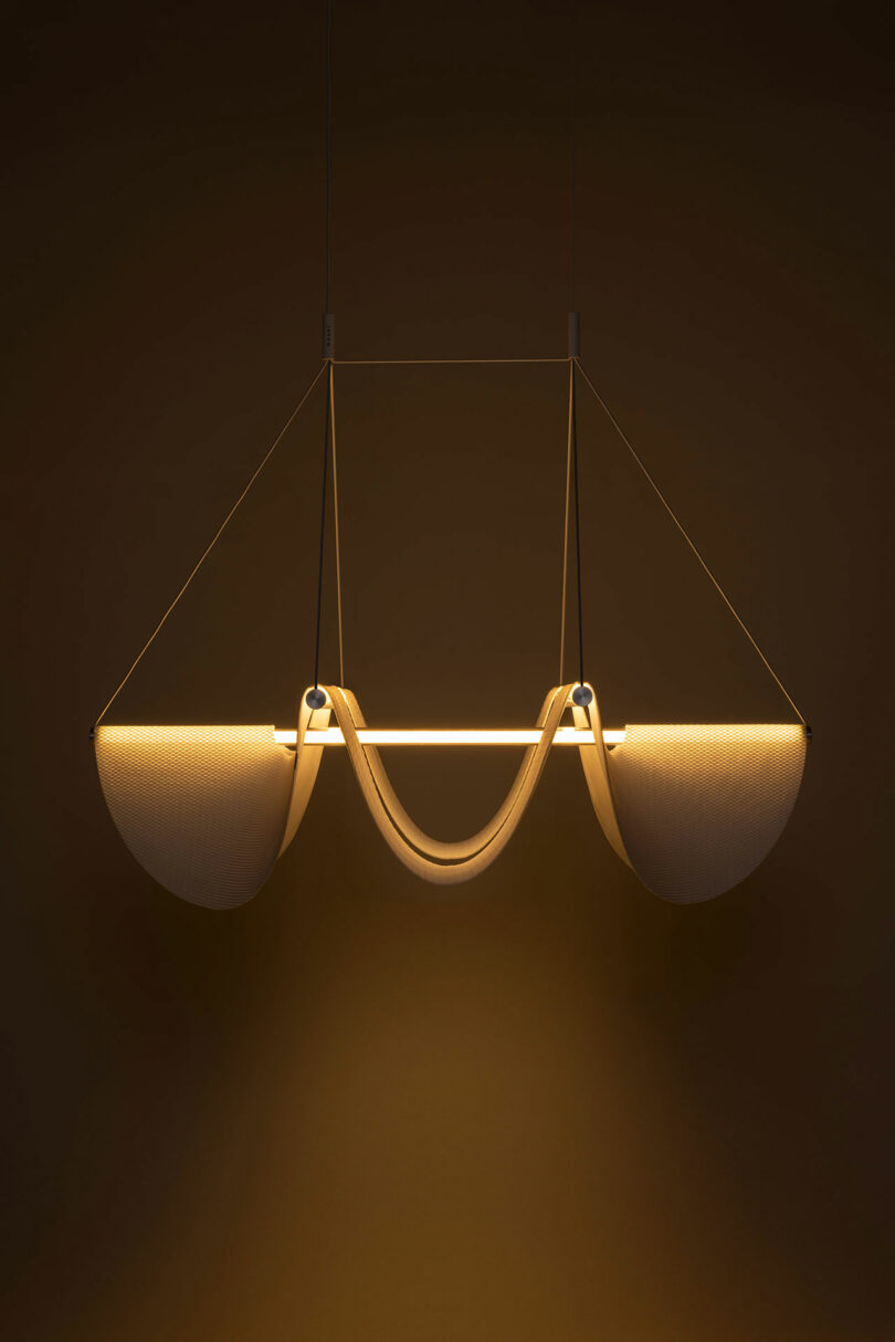 A Drape Light by Jamie Wolfond with a unique curved design, suspended from the ceiling and emitting a warm glow, set against a dark background.