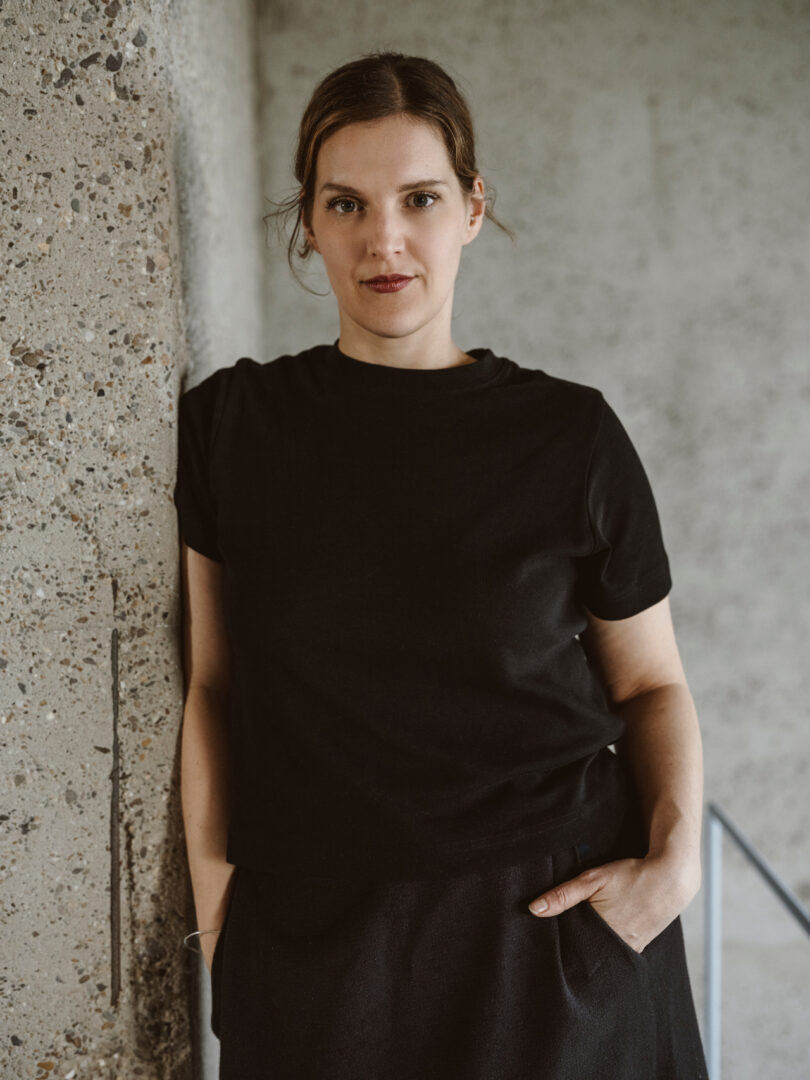 A person with short hair and a black t-shirt leans against a concrete wall with a neutral expression, with one hand in their pocket