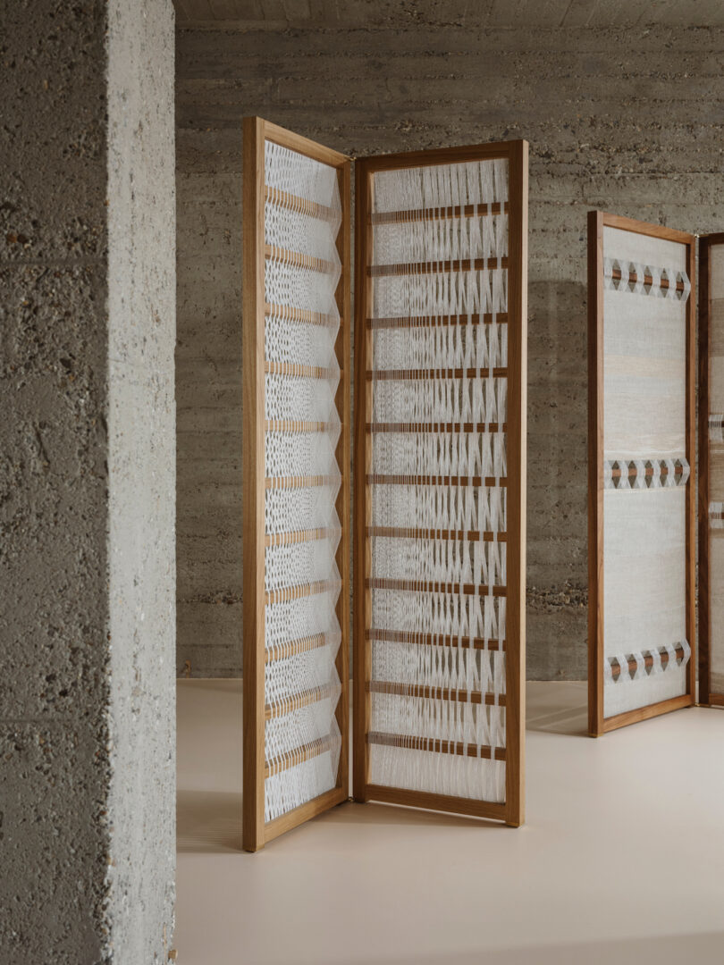 A wooden folding screen with a geometric, textured design stands against a concrete wall in a minimalistic room