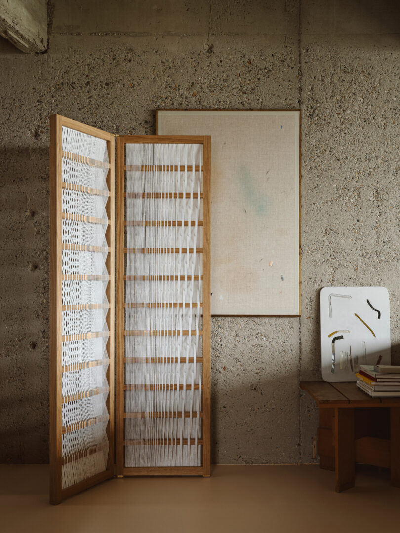 A minimalist room with concrete walls features a folding wooden room divider, a small table with books, a wooden chair, and a wall-mounted canvas