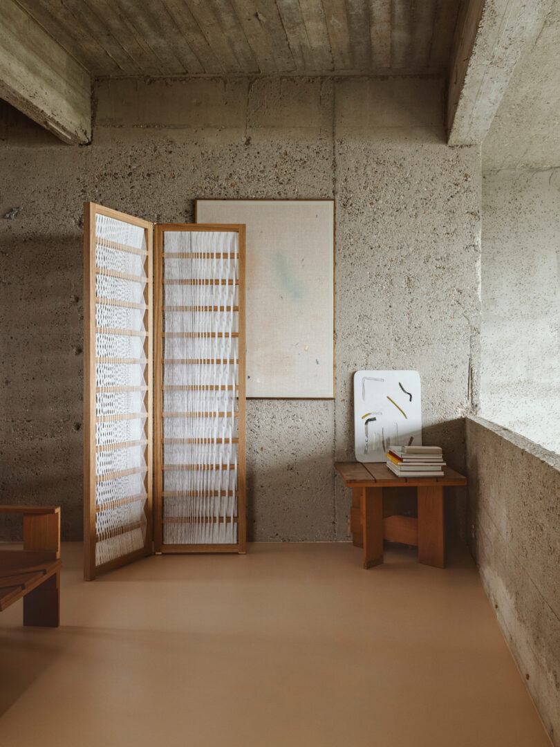 A minimalist room with concrete walls features a folding wooden room divider, a small table with books, a wooden chair, and a wall-mounted canvas