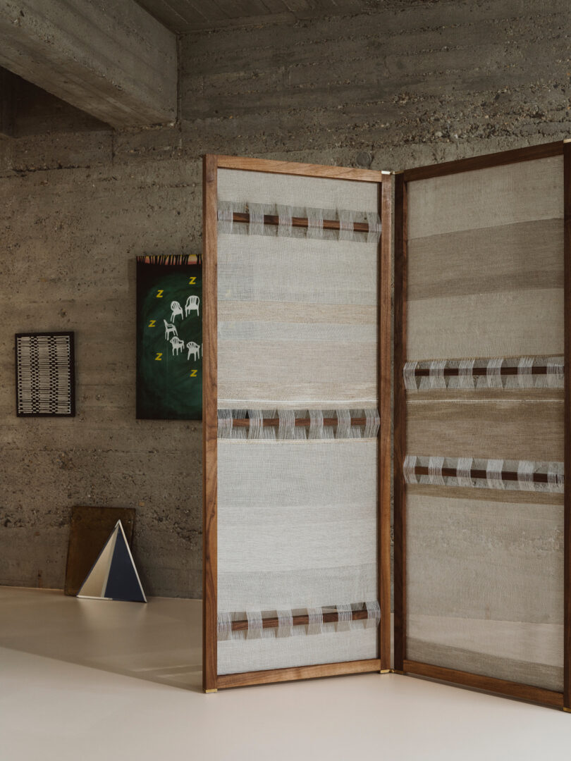 A room with concrete walls displaying a framed artwork, a wooden room divider with woven fabric, and a triangular object on the floor
