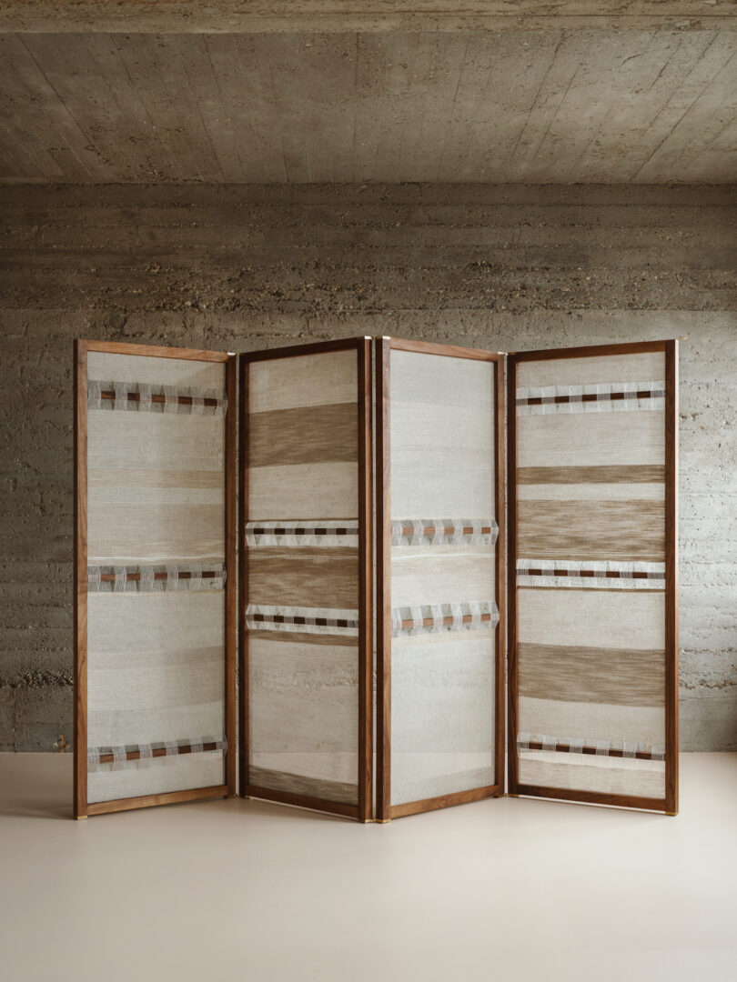 A four-panel room divider with a neutral colored geometric pattern stands against a concrete wall and a light-colored floor