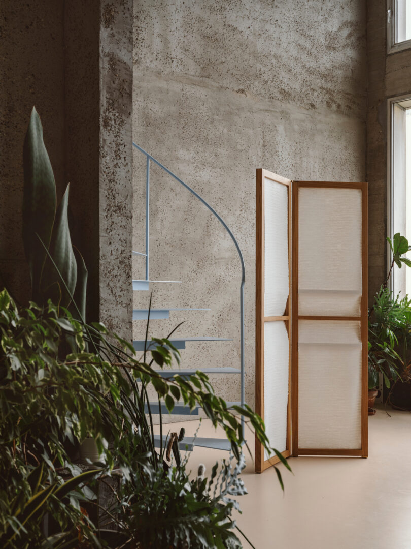 A cozy indoor space with a spiral staircase, various potted plants, and a wooden folding screen against a concrete wall
