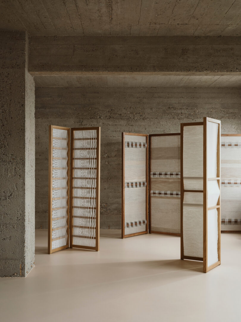 A minimalist room featuring tall, wooden-framed screens placed on a smooth, light-colored floor against textured concrete walls.
