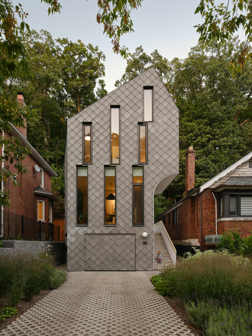 Modern, narrow, multi-story house with a unique, geometric façade, located between two traditional brick houses, with a stone pathway and greenery in the front.